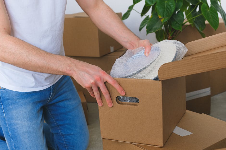 A person wearing a white T-shirt and blue jeans is packing a fragile item, specifically a large circular glass or ceramic plate, into a sturdy brown cardboard box. The person is holding the plate care