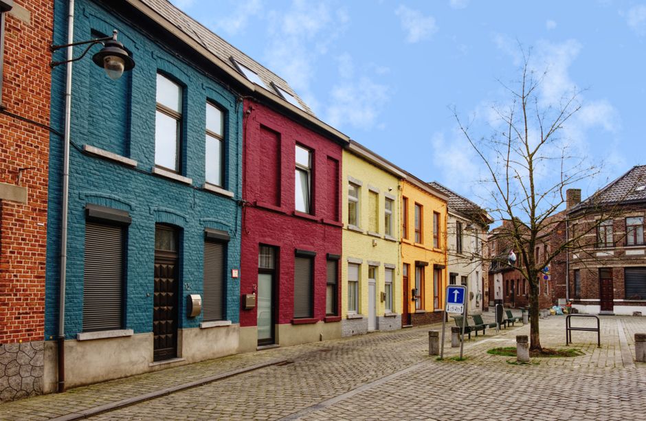 The image shows a row of historic, ornate townhouses with distinctive facades in a European city square, featuring a mix of brick and painted exteriors in red, brown, and white tones. Several flags ar